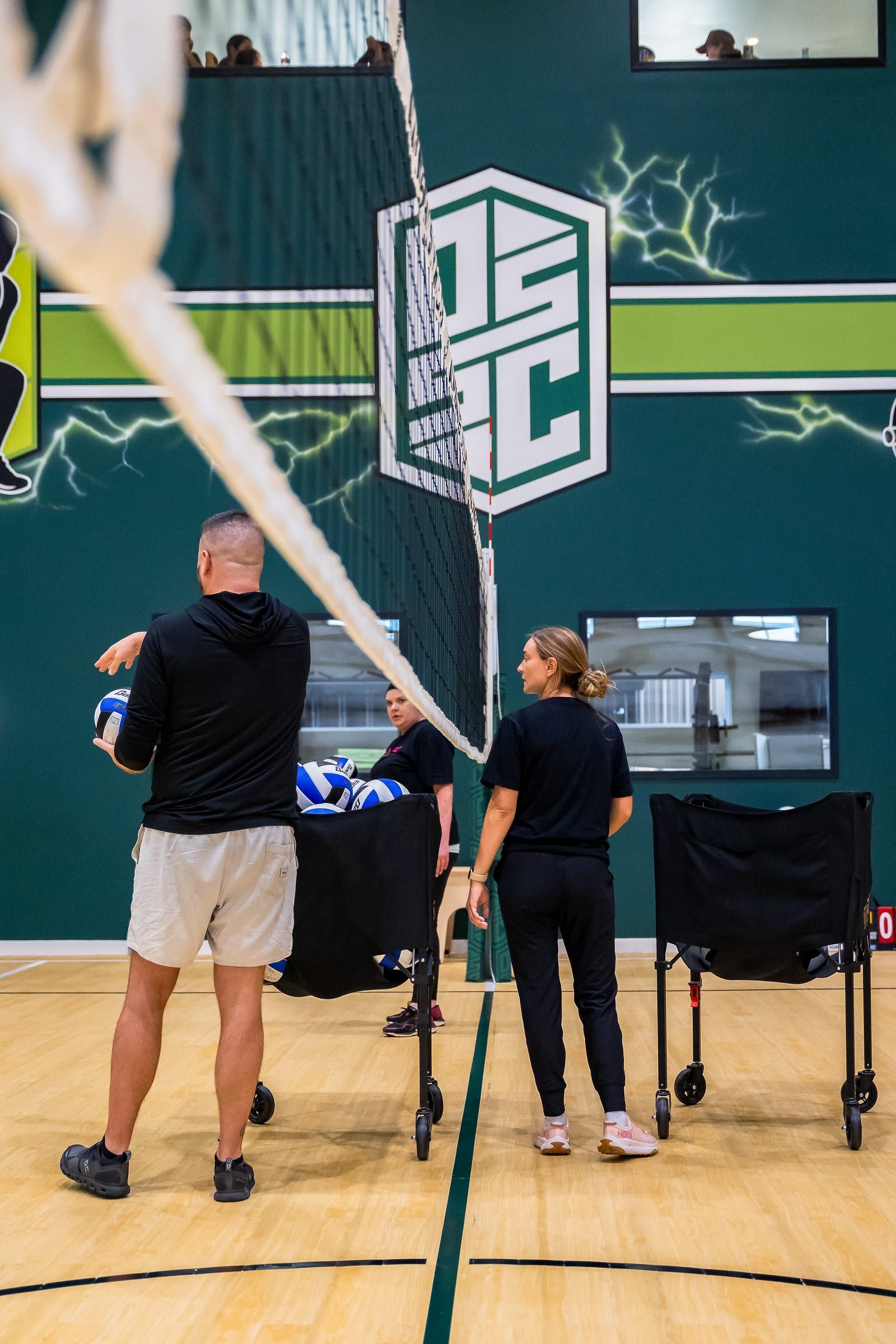 A coach and player talking at the net with a cart of volleyballs