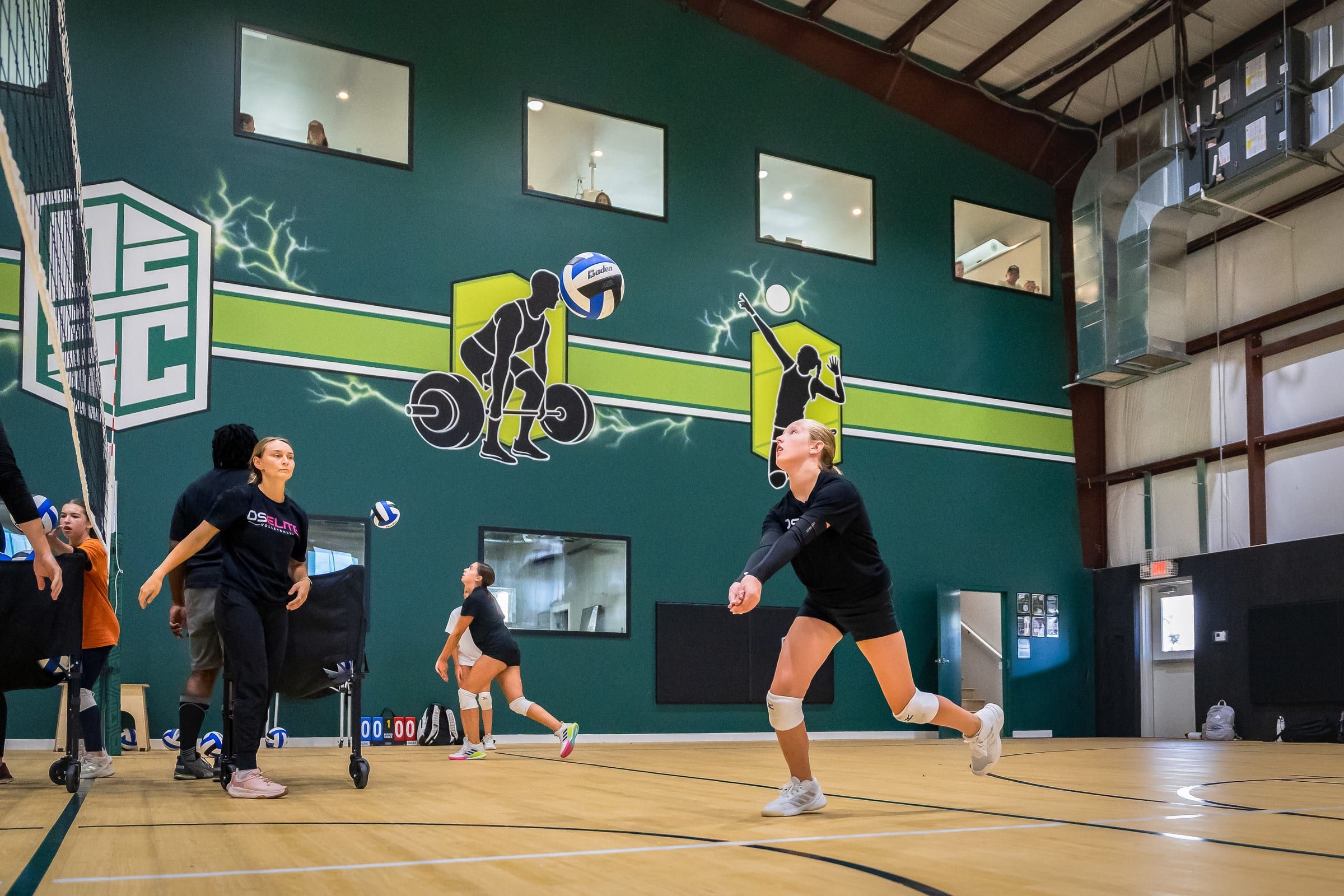A line of young volleyball players leaping at the net under the lights of the Warehouse, DSSC banners on the wall behind them