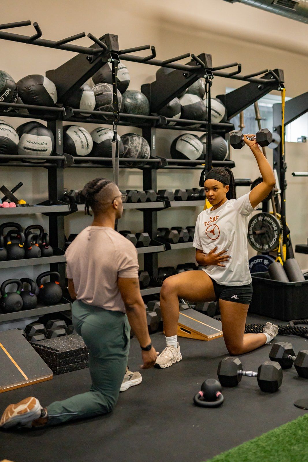 A Reach coach kneeling beside an athlete as she presses a dumbbell overhead, med balls racked behind them