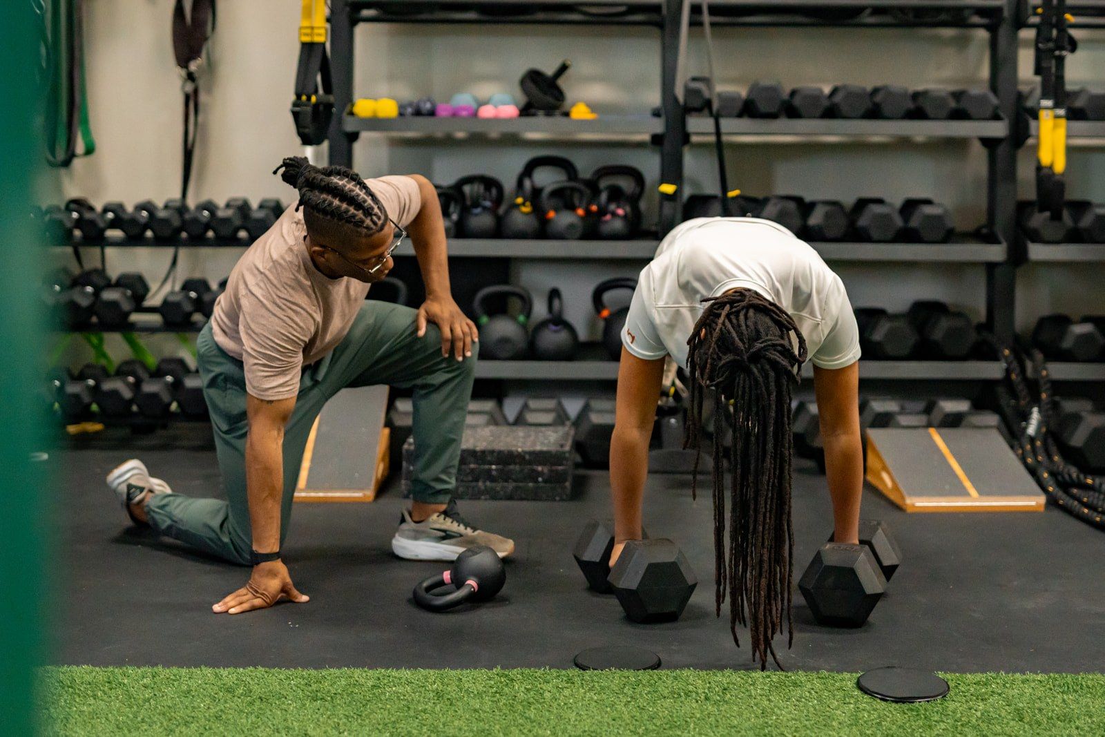A Reach coach and an athlete in a deep lunge stretch on the turf, kettlebells lined up behind them